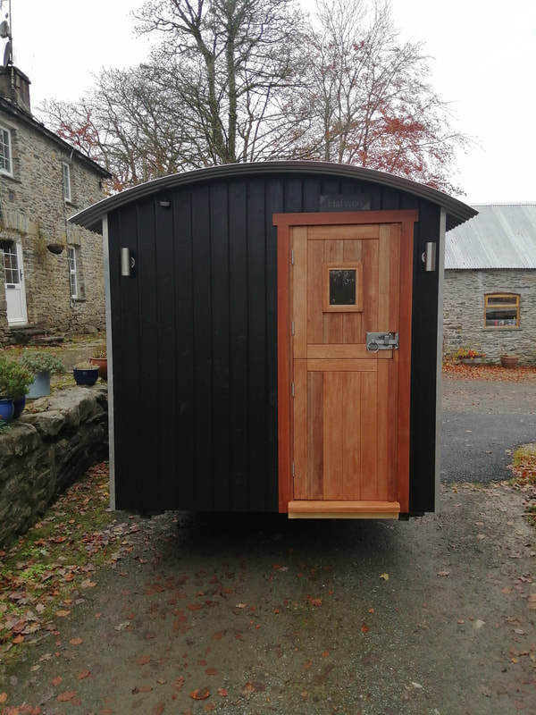 A black-clad shepherd's hut named Habren, with a timber door, parked in a stone-built farmyard.