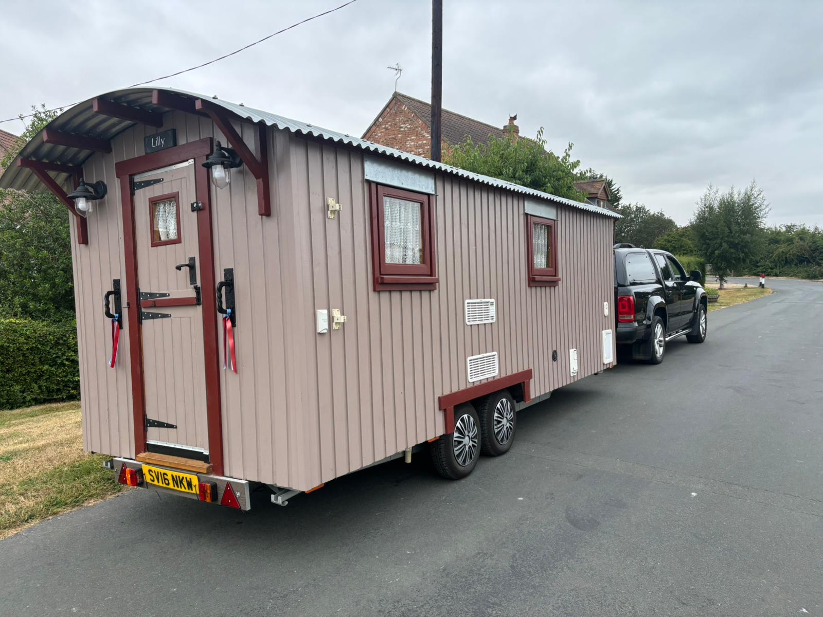 A dusky pink shepherd hut on a towable chassis on a village road.