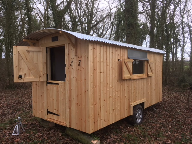 A pale timber shepherd's hut with its stable door open, standing among autumn leaves in woodland.