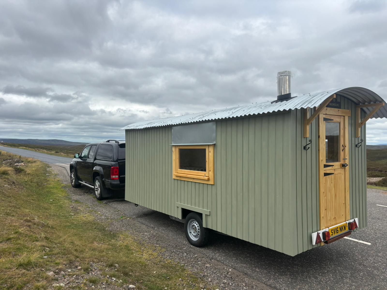 A sage-green shepherd's hut named Awelon, hitched to a pickup truck on a wild moorland road.