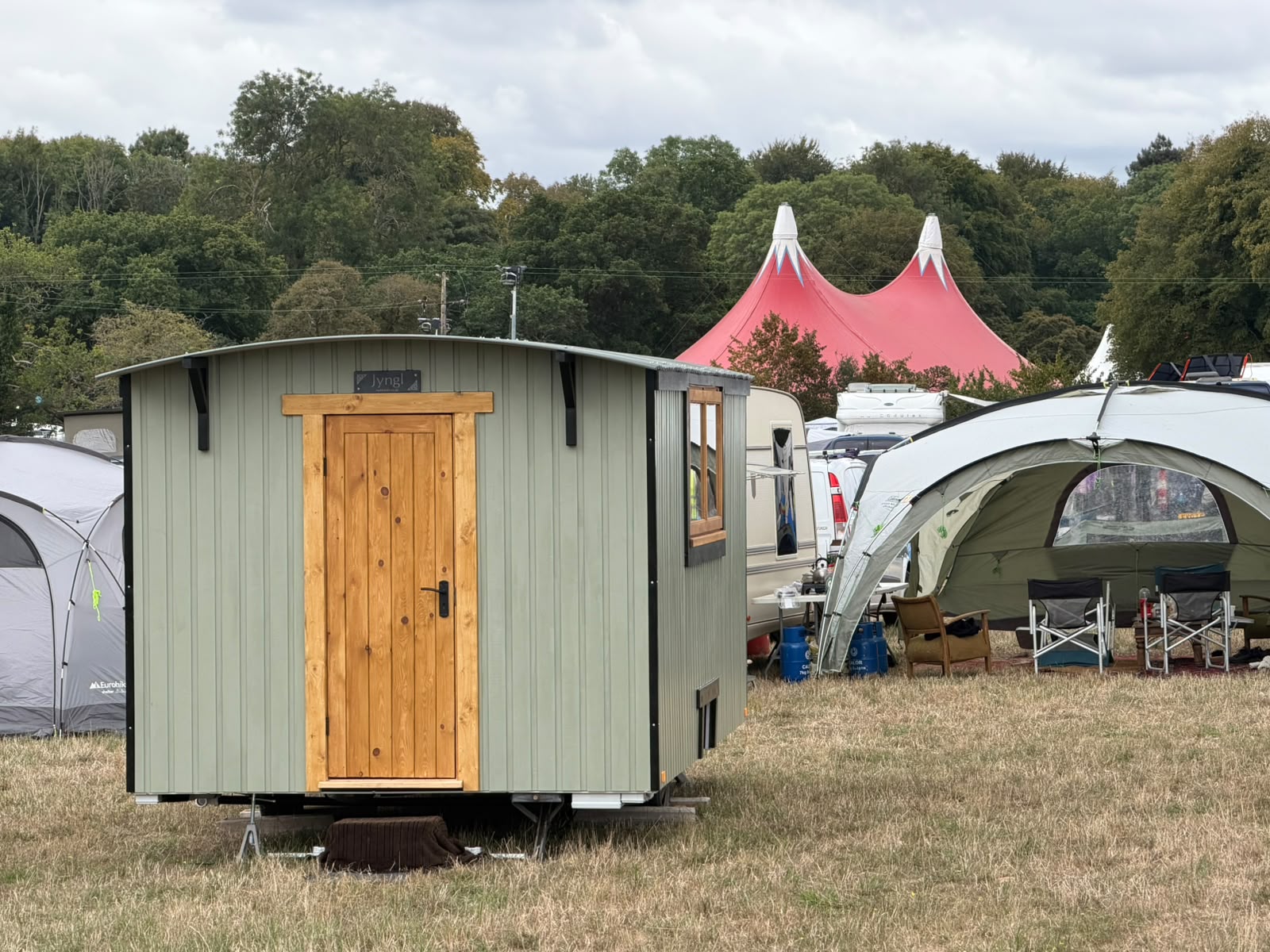 Jyngl, a sage-green shepherd's hut with a timber door, pitched at a festival campsite with a big top tent in the background.