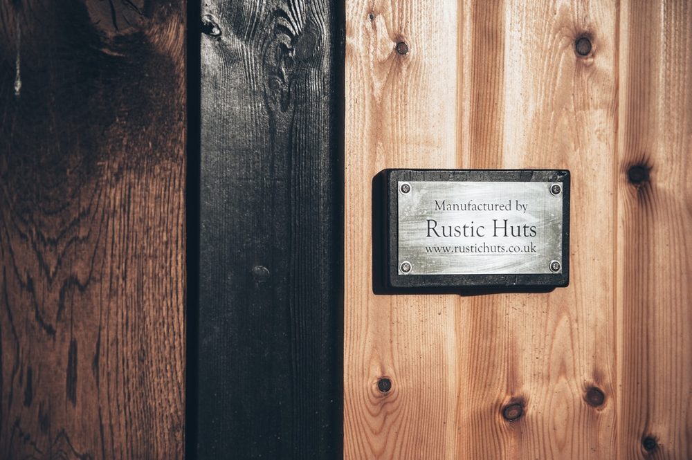 An engraved plaque reading Manufactured by Rustic Huts, mounted on a hut's timber cladding.