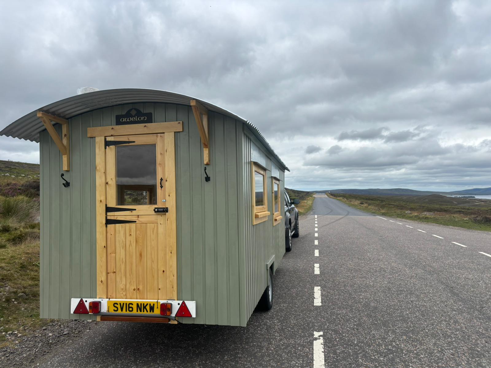 A green shepherd's hut on a towable chassis, hitched behind an ordinary car on a remote Scottish road.