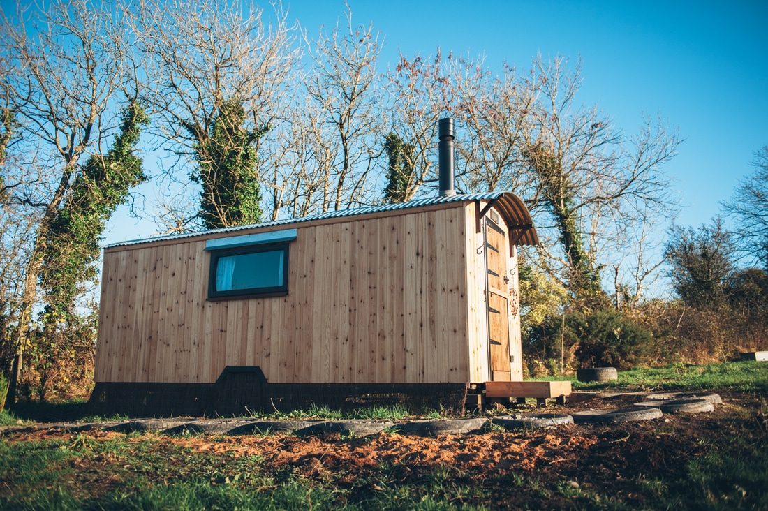 A handcrafted timber shepherd's hut with a curved roof, set among woodland, built by Rustic Huts in Carmarthenshire, Wales.